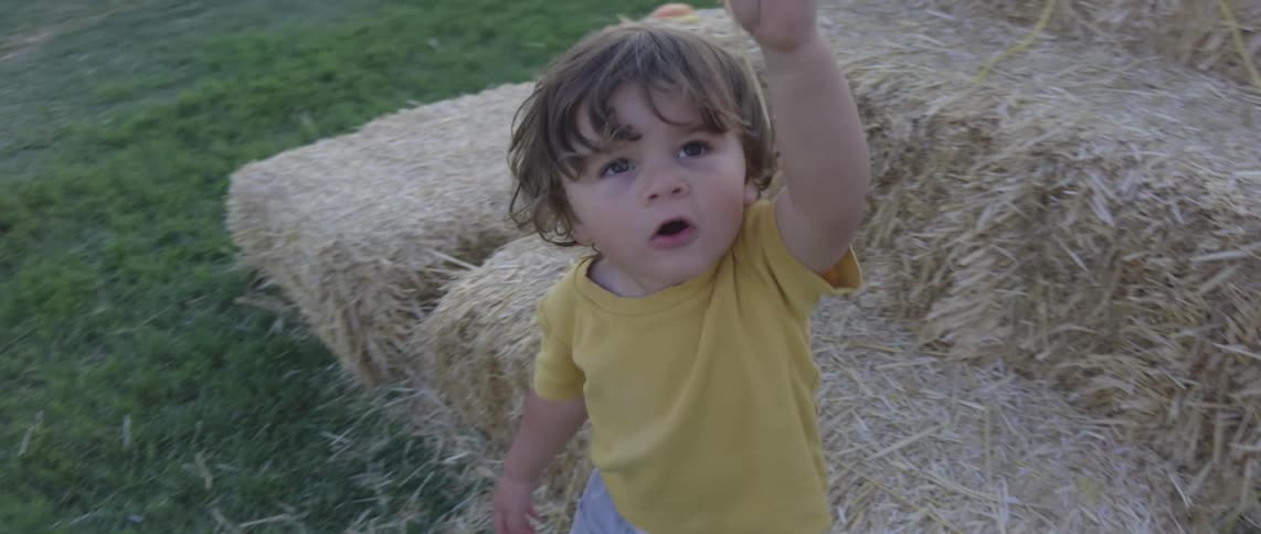 A toddler in a yellow shirt reaches up and plays with straw near hay bales and pumpkins