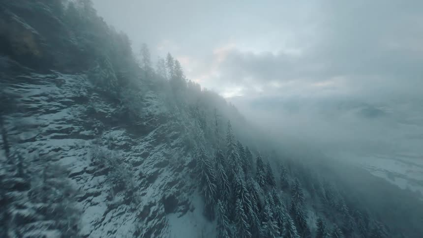 A drone captures a snowy mountainside its trees dusted in white as fog blankets the valley below