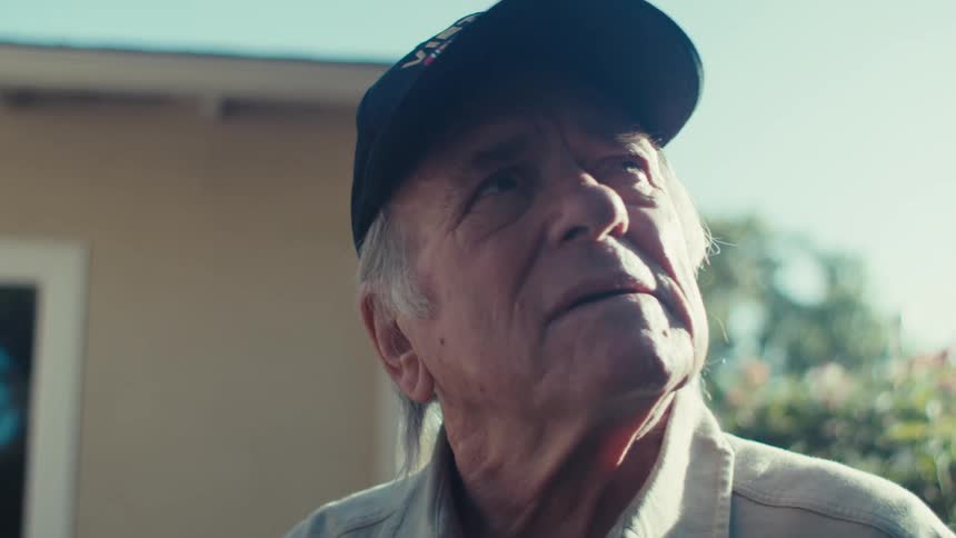 Elderly man wearing a veteran cap stands in the house yard while looking around on a sunny day