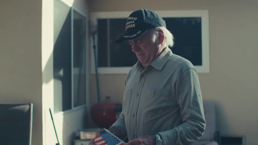 An old man stands outside his house looking sad as he holds and looks at a small american flag