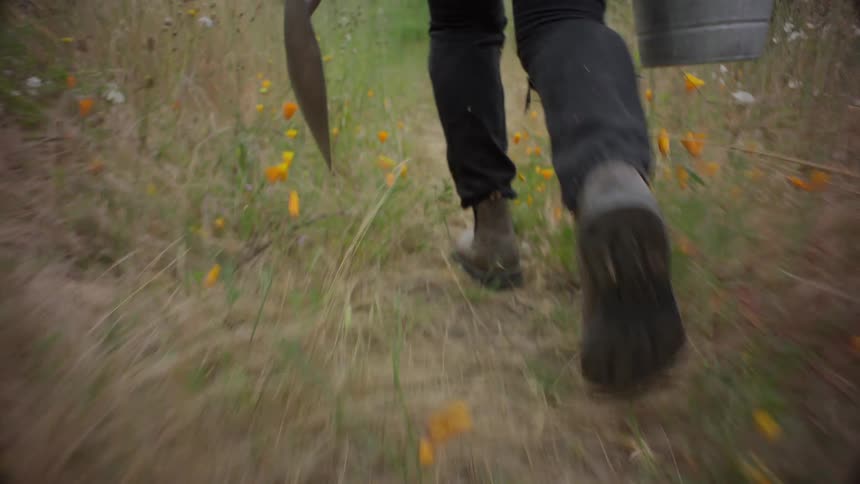 Close up of the legs of a person carrying a bucket and a shovel through a field