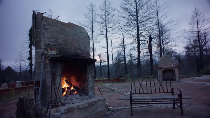 A fireplace with a roaring fire surrounded by the ruins of a house and a metal bench