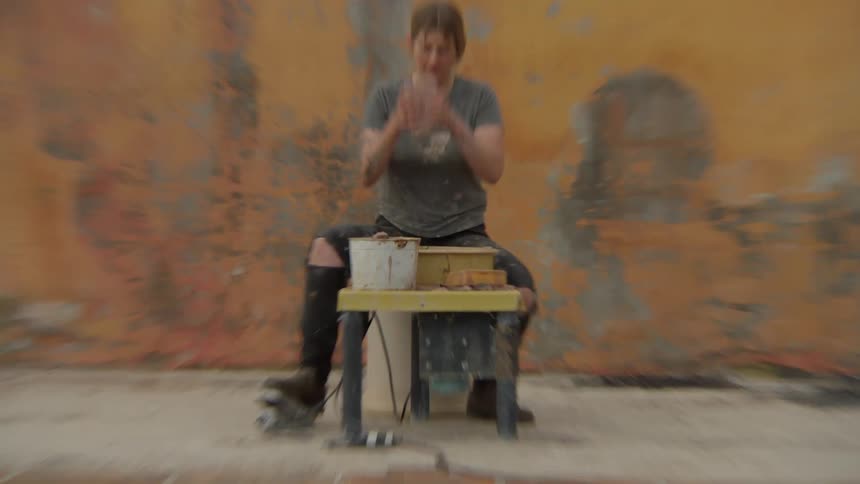 A woman places a piece of clay on a turning table in front of a weathered wall