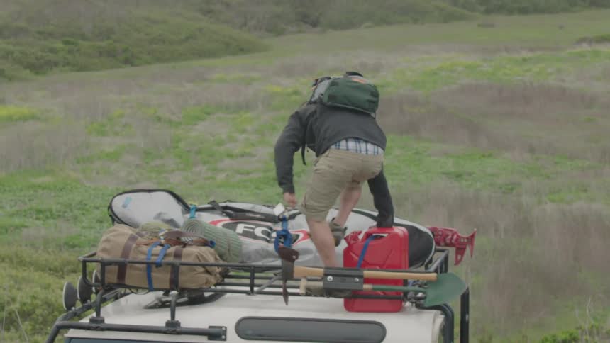 A man with a backpack climbs onto a jeep loaded with gear and stows his backpack