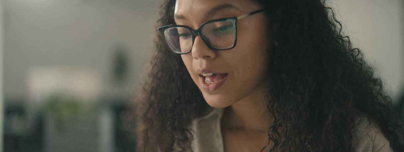 Woman with curly hair glasses and a brown shirt talks to a person in the office