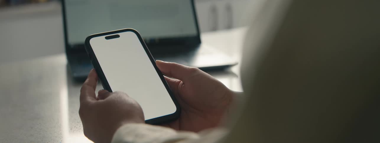 A person holds a smartphone with a blank screen with a laptop in the background