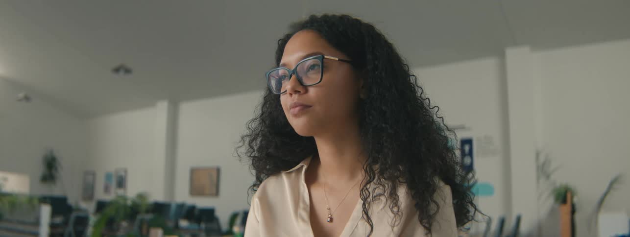A woman with glasses and curly hair looks thoughtfully to the side
