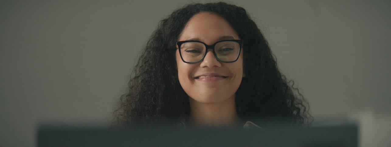 A woman with glasses and curly hair laughs while looking at her computer
