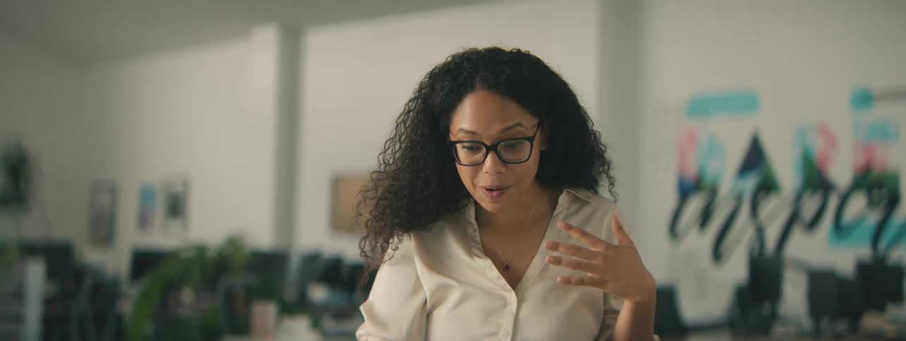 A happy woman with curly hair gestures while talking on a video call through a laptop in the office
