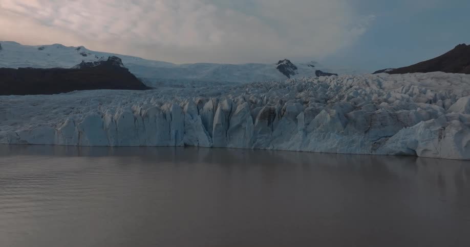 Shore side ocean water reflects a glacier under an overcast sky