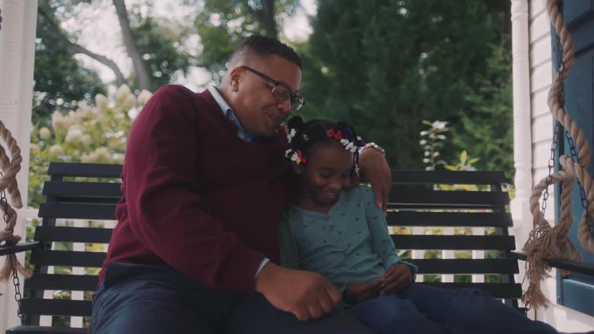 A father and daughter sit on a hanging chair on the porch talking