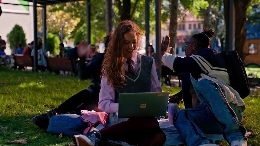 A group of students sit on the grass at the college campus greeting another student who joins them