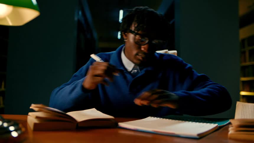 A student in a blue sweater studies and takes notes in a library at night