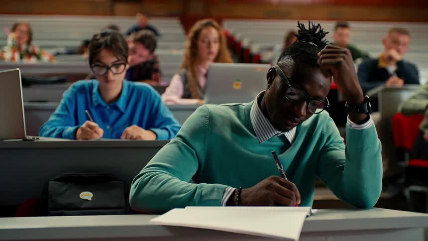 Students in a university classroom writing in notebooks and typing on a laptop