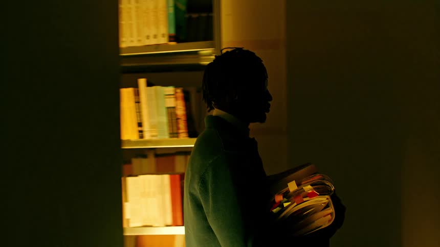 A young man picks up books from the shelves in a dimly lit library
