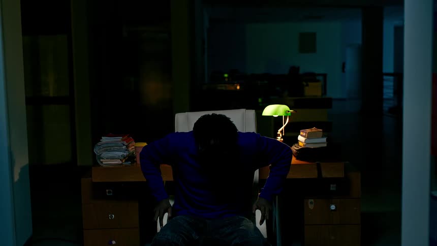 A young man sits at a desk in the library at night stands up tired and walks through the shelves