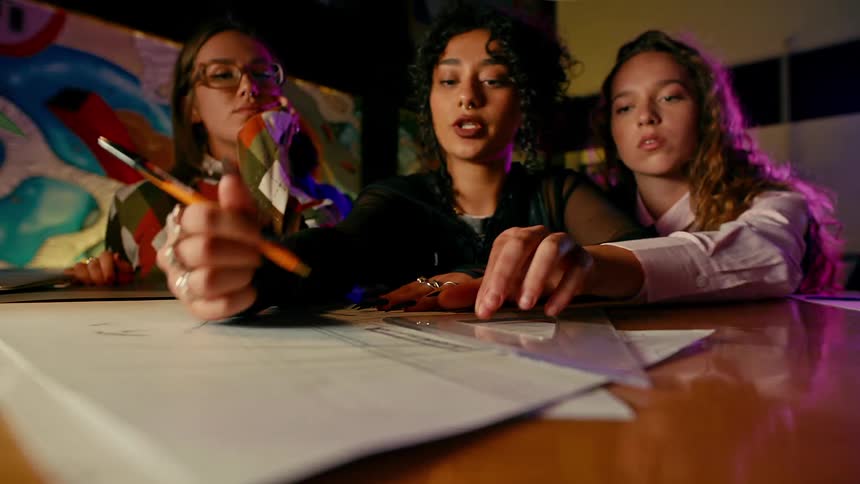 Three college girls sitting with one of them with a pencil drawing on a sheet of paper on the table