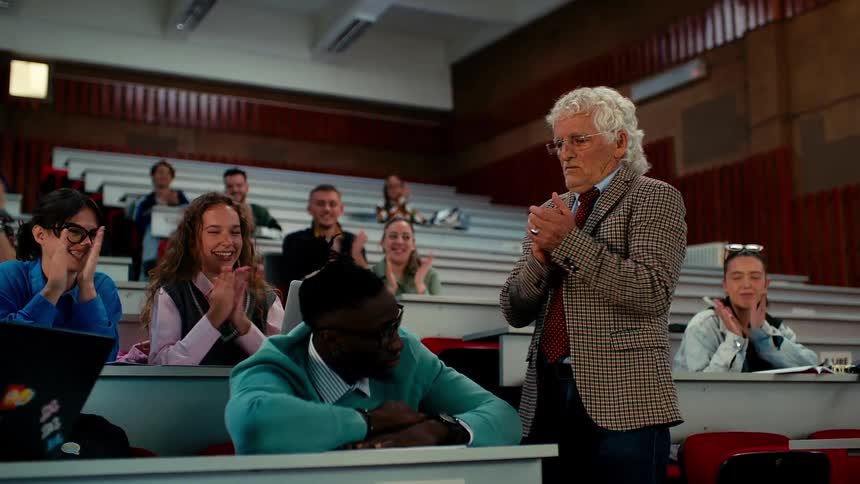 A professor and students clap cheerfully to wake up a sleeping classmate in a college classroom