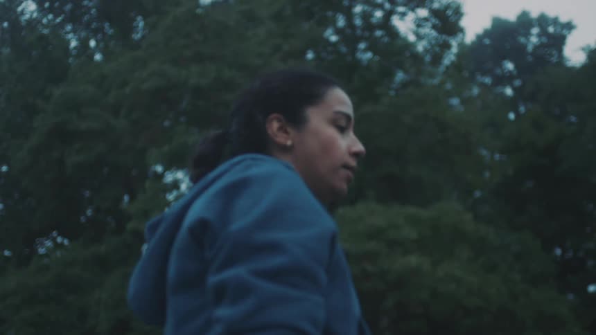 A woman wearing a green zipup hoodie takes a deep breath as she walks past green trees