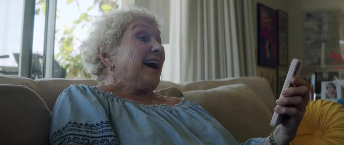 A happy elderly woman laughs while looking at her smartphone sitting on the couch at home
