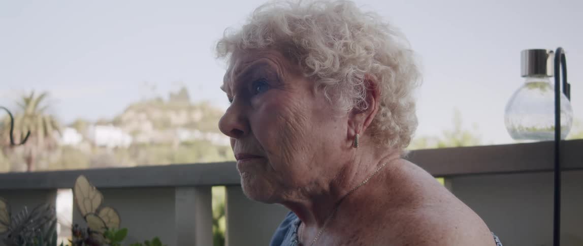 An elderly woman sitting on a porch reading on a sunny day