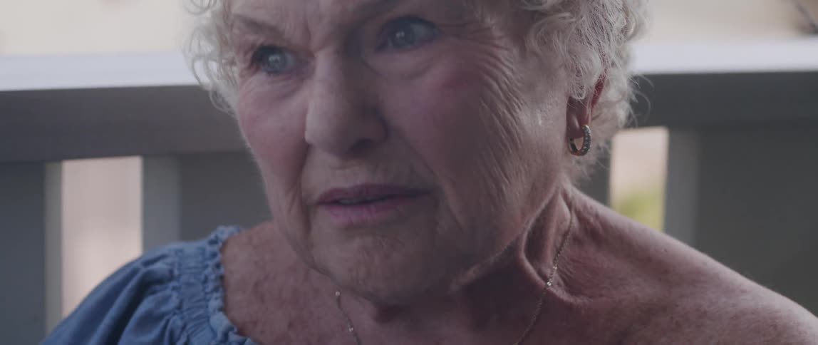 An old woman rubs her hands together while smiling and talking on a porch