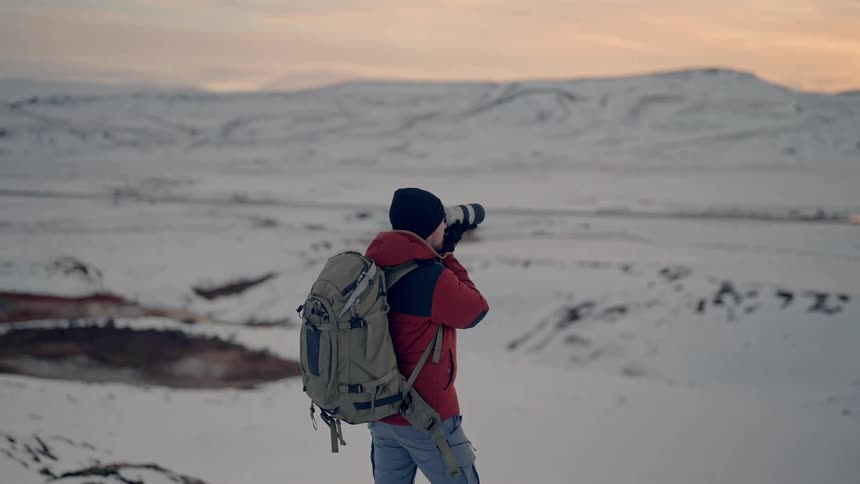 A man hikes up on snow covered elevated land and takes a photograph of a mountain range