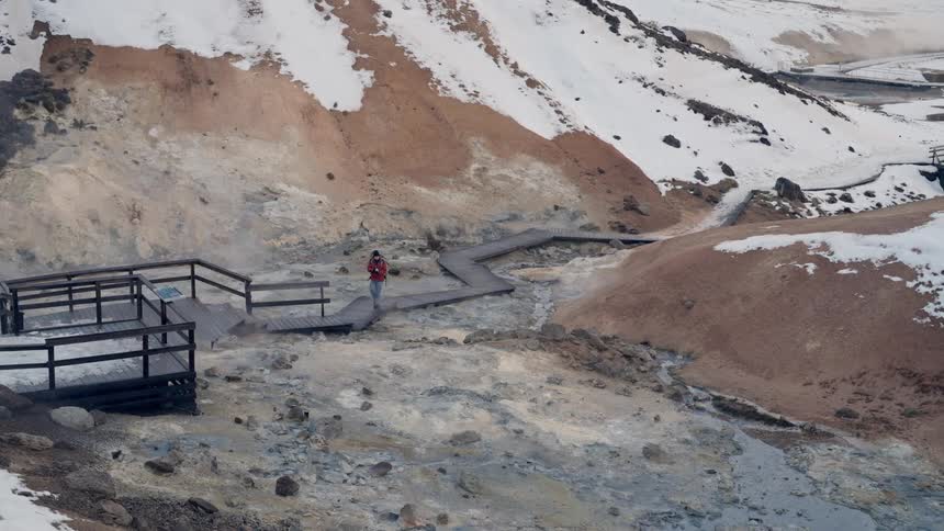 Male photographer hiking along the wooden path over the terrain near a mountain stops to take a picture on a winter day