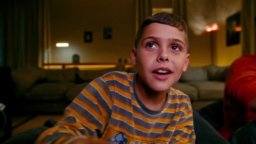 A young boy sits next to his brother while talking and playing a videogame