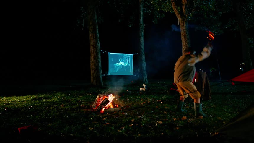 A young boy swings a torch near a campfire while a film is projected onto a sheet hung between two trees at a campsite at night