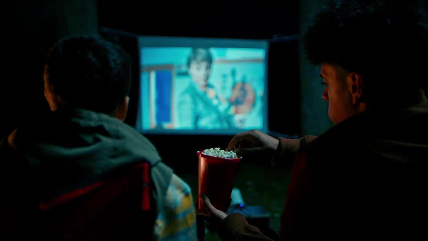 Two people are watching a small projector screen and eating popcorn