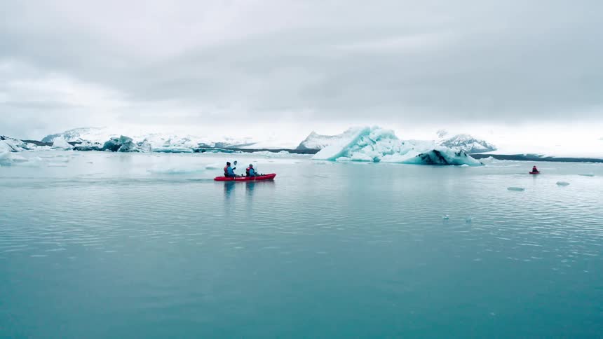 Two kayakers paddle through a serene glacial lagoon dwarfed by the towering icebergs that dot the horizon