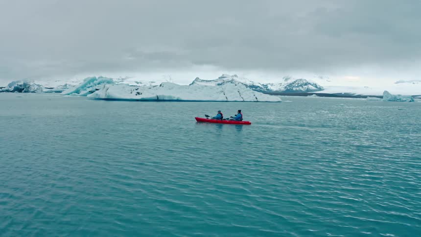 Two kayakers paddle through a serene glacial lagoon surrounded by majestic icebergs and snowcapped mountains