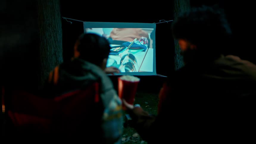 Two friends at a campsite watching a movie and eating popcorn
