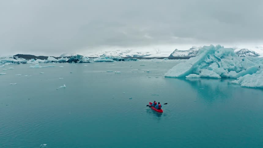 Two people paddle a red boat into a cove with floating ice and a snow and ice clogged shoreline