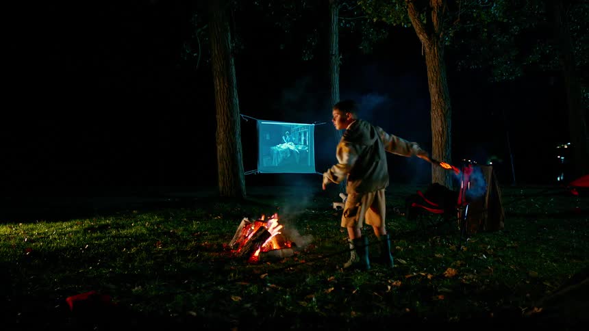 A boy stands next to a campfire swinging around a roasted marshmallow on a stick