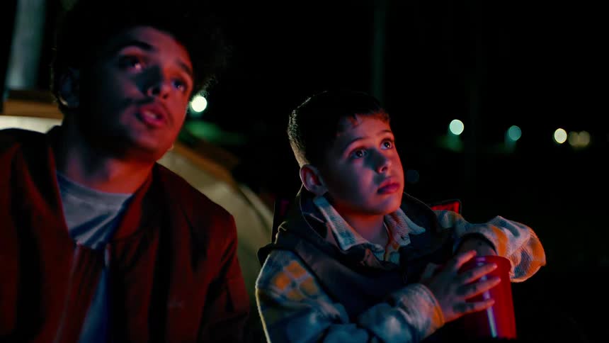At night two boys sit and watch something with looks of wonder on their faces the younger boy eating from a red container