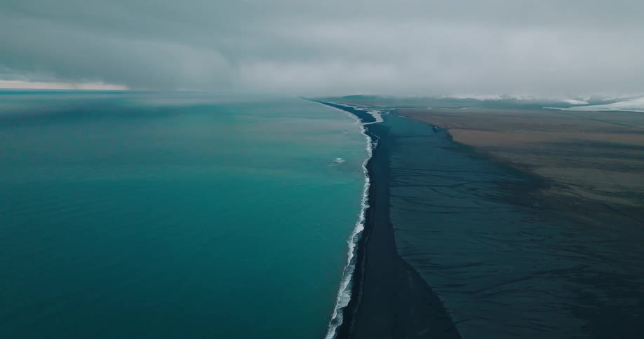 A dramatic aerial view of a black sand beach meeting the turquoise ocean under dark clouds