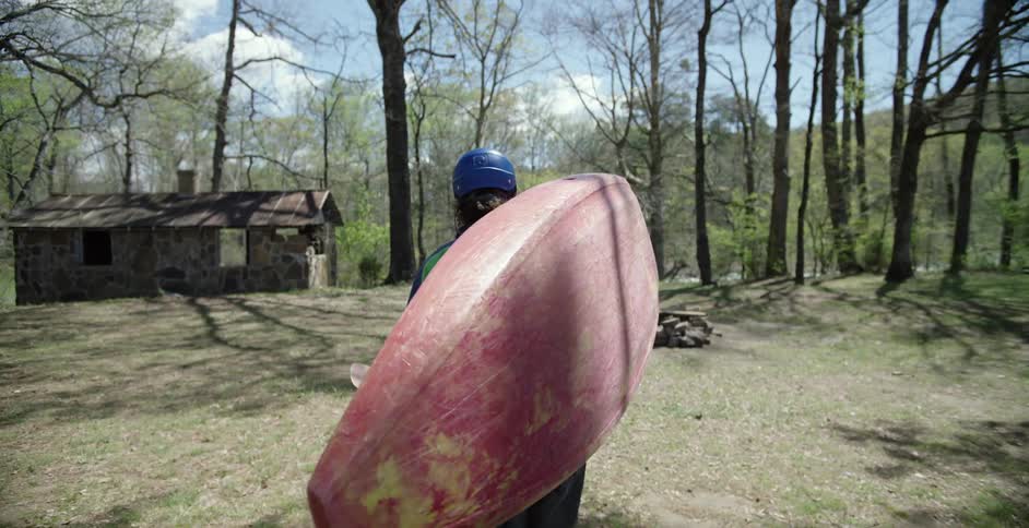 A person is walking in a wooded area with his kayak over his shoulder
