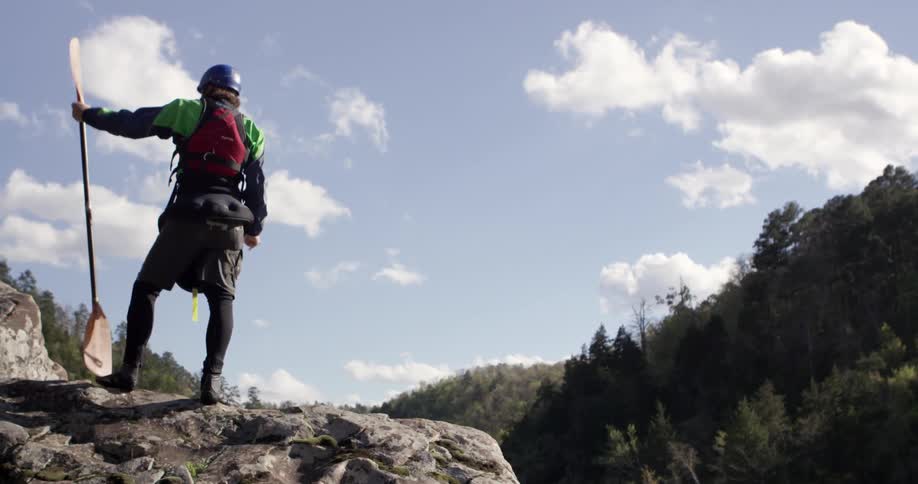 Kayaker standing atop a rocky hill holding his paddle on a partly cloudy day