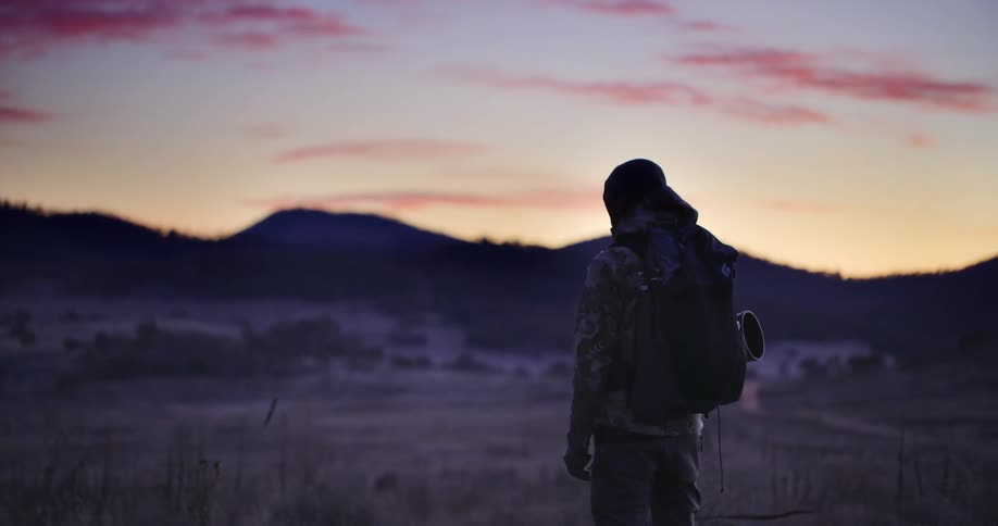 Silhouette of a person with a backpack in a field near mountains under a twilight sunset sky
