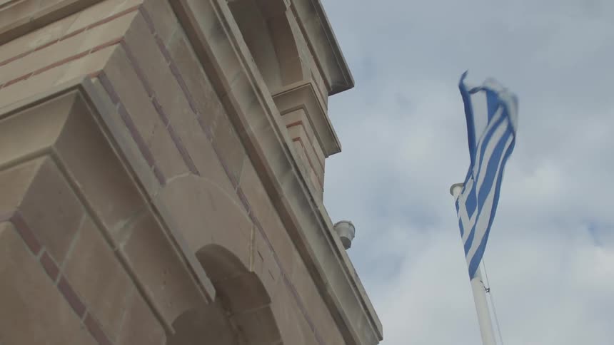 A greek flag waves in the wind attached to a historic building