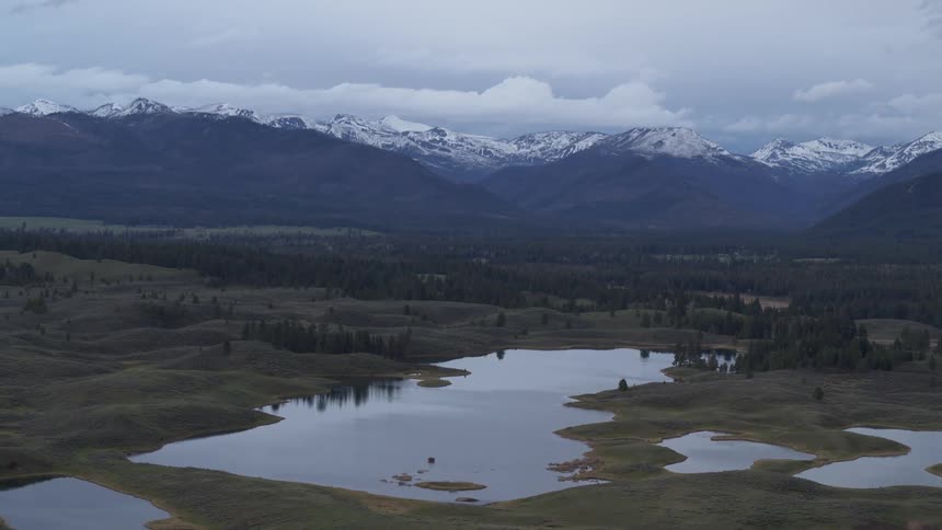 Tranquil landscape with small lakes surrounded by grassy areas with majestic mountains partially covered in snow in the background under a cloudy sky