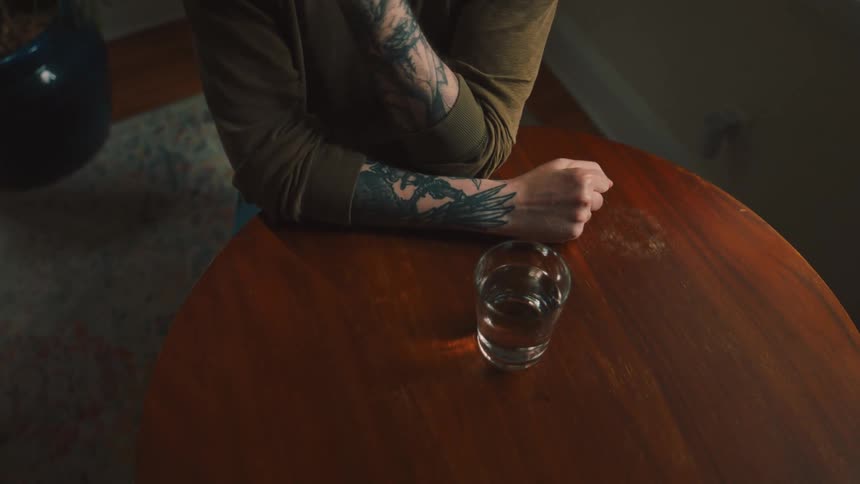 A pair of tattooed arms rest on a table with a glass of water