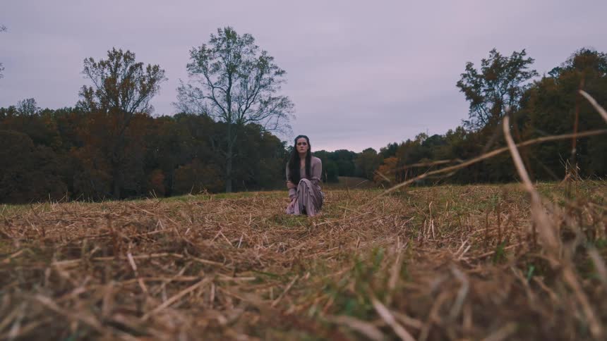 Woman in a purple dress crouching in a field surrounded by trees