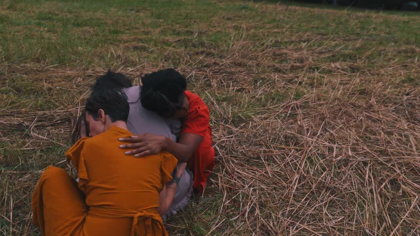 Three women in different colored dresses crouching together in a field