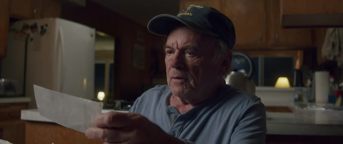An elderly man sits in his kitchen laughing as he opens a letter