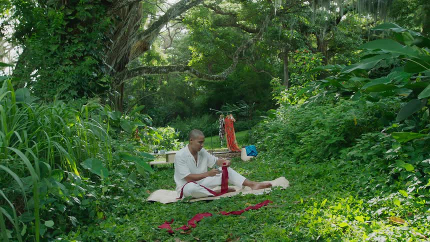 Man sitting on grass and an environment with plants and trees holding a red fabric