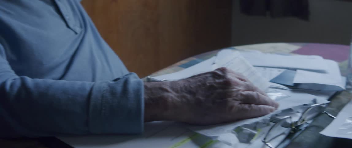 An elderly man looks sad as he reads sits in the kitchen with his hand over a letter