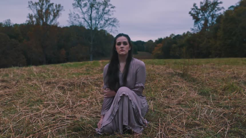 A woman in a dress sits in a field of dry grass looking at the camera